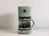 A green coffee maker with a glass carafe filled with coffee, placed on a marble countertop against a white wall. Náhled obrázku produktu