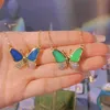 Zwei goldene Ketten mit Schmetterlingsanhängern, einer mit blauen Flügeln und der andere mit grünen Flügeln, beide mit kristallbesetzten Körpern, in einer Hand gehalten Náhľad obrázku produktu