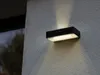 A black outdoor wall light with a motion sensor is mounted on a textured gray wall. Produktbild-Vorschau
