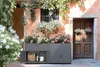 A gray planter box with storage is placed in front of a building with a wooden door and a window, surrounded by plants and flowers. Produktbild-Vorschau