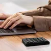 A person's hands typing on a laptop with a small black numeric keypad on the desk in front of them. Podgląd zdjęcia produktu