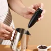A person is holding a black electric milk frother over a stainless steel pitcher, with coffee beans in a small white dish in the foreground. Produktbild-Vorschau