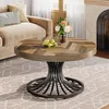 A round wooden coffee table with a metal base is placed on a rug in a living room setting, next to a sofa and a fireplace. Voir les images du produit
