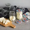 Several glass jars filled with cubed tofu and some empty jars are arranged on a surface, with a cutting board holding more tofu in the foreground. Produktbild-Vorschau
