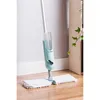 A spray mop with two cleaning pads attached to the base, standing on a wooden floor next to a wall and a plant. Náhled obrázku produktu