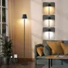 A floor lamp is shown in a living room setting next to a sofa and a window. Produktbild-Vorschau