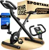 A black exercise bike is displayed on a light beige circular platform against a white background. A cardboard box with the brand name and product features is behind the bike. Produktbild-Vorschau