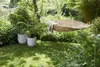 A serene garden scene with a woven hammock strung between trees, two potted plants on a lush green lawn, and dense foliage in the background. Náhľad obrázku produktu