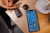 A person's hand is holding car keys with a tracker attached, next to a smartphone displaying a map application. Podgląd zdjęcia produktu