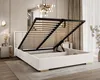 A white upholstered bed frame with its storage compartment open, revealing the slatted base and metal frame. The bed is in a well-lit, modern bedroom setting with decorative elements. Produktbild-Vorschau
