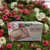A product box with a floral design, featuring a pink handbag-shaped mug set with a saucer and spoon, placed on a bed of green moss and pink and white roses. Produktbild-Vorschau