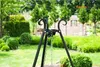 A black metal tripod stand with chains is set up in a garden with lush green bushes and trees in the background. Náhled obrázku produktu
