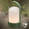 A green LED table lamp with a frosted white shade is shown outdoors in the rain. The lamp is lit with a warm white light. An IP44 rating symbol is visible in the lower right corner. Anteprima dell’immagine prodotto