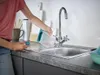 A person is pouring water from a clear plastic container with a teal handle into a kitchen sink. Anteprima dell’immagine prodotto