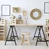 A home office or craft room setup with a wooden desk supported by black trestle legs, a wooden stool, and various storage crates and decorative items on the wall. Podgląd zdjęcia produktu