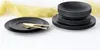 A set of black stoneware plates and bowls are arranged on a white surface with a yellow fabric draped in the background. Náhľad obrázku produktu