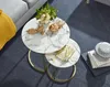 A set of nesting coffee tables with marble-look tops and gold metal frames, styled with decorative items and a throw blanket. Produktbild-Vorschau