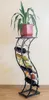 A black metal wine rack with bottles of wine and a potted plant on top, set against a light-colored wall. Anteprima dell’immagine prodotto