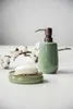 A green ceramic soap dispenser and a soap dish with a bar of soap are placed on a white surface next to a branch with cotton bolls. Produktbild-Vorschau