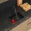 A modern kitchen sink with a black granite sink, a black faucet with water running, and three red tomatoes inside the sink. The countertop is black, and the cabinets below are made of light brown wood. A wooden cutting board is visible on the countertop. Podgląd zdjęcia produktu