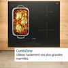 A black induction hob with a roasting pan containing food is shown on a wooden surface. Text overlays describe a feature. Voir les images du produit