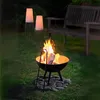 A fire pit with flames and logs is set up on a grassy lawn at night, with two illuminated lamps and a wooden bench in the background. Produktbild-Vorschau