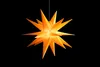 A lit, orange, star-shaped lantern hangs against a black background. Produktbild-Vorschau