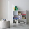 A white bookshelf with various items on its shelves, next to a round wooden table with a magazine on it. Produktbild-Vorschau