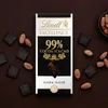 A Lindt Excellence 99% Cocoa dark chocolate bar is displayed on a dark brown background, surrounded by loose cocoa beans and individual chocolate squares. Produktbild-Vorschau