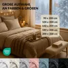 A cozy bedroom scene with a bed made up with bedding, a knitted blanket, and a mug of hot drink, set against a snowy window view. Various color swatches of bedding are shown at the bottom. Produktbild-Vorschau