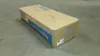 A cardboard box containing a product, likely toner, is placed on a concrete floor. Voir les images du produit