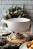A fondue set is displayed on a wooden table, with a decorative wreath in the background. Produktbild-Vorschau