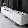 A white media console with silver accents and metallic cylindrical legs, positioned in front of a large black screen and a dark background. Voir les images du produit