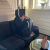 A person wearing a black hooded blanket sits on a dark couch and reads a book. Náhled obrázku produktu