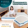 A woman sitting on a mattress topper on a bed, with marketing text and logos. Produktbild-Vorschau