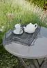 A metal tray with a teapot and two cups sits on a round outdoor table, surrounded by greenery. Produktbild-Vorschau