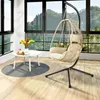 A hanging egg chair with a cushion is set up on a wooden floor next to a small white table with a vase of flowers and some tea set. Produktbild-Vorschau
