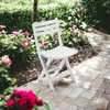 A white folding chair sits on a paved patio surrounded by lush green foliage and vibrant pink flowers. Náhled obrázku produktu