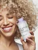 A woman with curly blonde hair smiles as she holds a bottle of Olaplex No.5P Blonde Enhancer Toning Conditioner. Náhled obrázku produktu