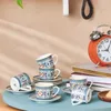 A set of coffee cups and saucers with a decorative pattern, arranged on a light blue surface, with an orange alarm clock and books in the background. Produktbild-Vorschau