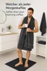 A man in a bathroom is wrapped in a large dark gray towel, holding a white mug and smiling. Produktbild-Vorschau