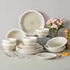 A collection of stoneware dinnerware is arranged on a flat surface against a light gray background, with a vase of pink flowers and a wooden pepper grinder to the right. Produktbild-Vorschau