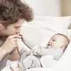 A father is kissing his baby's foot while the baby lies in a white bassinet. Anteprima dell’immagine prodotto