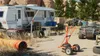 A campsite scene with a trailer, generator, ATVs, and various equipment scattered around. Náhľad obrázku produktu