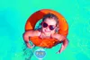 A young girl wearing heart-shaped sunglasses floats in a pool wearing an orange inflatable ring, with a thermometer floating nearby. Náhľad obrázku produktu