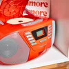 A close-up of an orange Blaupunkt boombox with its CD lid open, showing a CD inside. The background is a shelf with some text that reads 'more more'. Produktbild-Vorschau