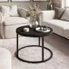 A round coffee table with a dark concrete top and black metal legs is placed on a plush white rug in a living room setting. It is surrounded by a cream-colored armchair and a sectional sofa. Náhľad obrázku produktu