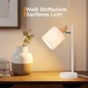 A desk lamp with a fabric shade and wooden accents is placed on a wooden desk next to a vase with a plant and a glass of water. Produktbild-Vorschau