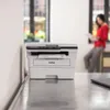 A Brother printer is shown on a desk in an office setting, with a woman in the background. Produktbild-Vorschau