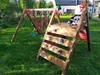A wooden playground set with swings, a climbing wall, and a slide, set up in a grassy backyard with a house in the background. Anteprima dell’immagine prodotto